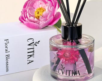 Può includere: Una bottiglia diffusore in vetro trasparente con un tappo nero e canne nere. La bottiglia è riempita con un liquido viola e contiene fiori rosa e bianchi. La bottiglia è etichettata "CVITKA" e la scatola è etichettata "Floral Blossom CVITKA".