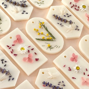 Könnte beinhalten: Eine Sammlung weißer Wachs-Melts in Anhängerform, verziert mit getrockneten Blumen und Kräutern. Die Melts zeigen verschiedene Blumenarrangements, darunter Lavendel, Mimosen, Gänseblümchen und rosa Hortensien. Jeder Melt hat oben ein kleines Loch.