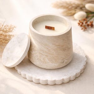Può includere: Una candela beige marmorizzata in un contenitore rotondo e strutturato con uno stoppino di legno. La candela è appoggiata su un vassoio bianco con bordo smerlato, con il coperchio accanto. Piante secche sullo sfondo.