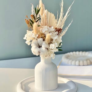 Könnte beinhalten: Eine weiße Keramikvase mit geripptem Design, gefüllt mit einem Trockenblumenarrangement. Das Arrangement umfasst weiße Hortensien, Baumwollknäuel, getrocknete Gräser und Pampasgras.