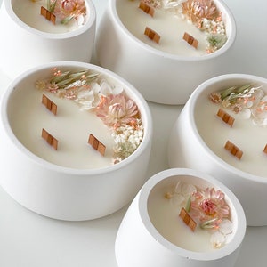 May include: Five white ceramic candle holders with a rounded, bowl-like shape. Each holder features a different arrangement of dried flowers and greenery, including white, pink, and green tones. The candles have wooden wicks.