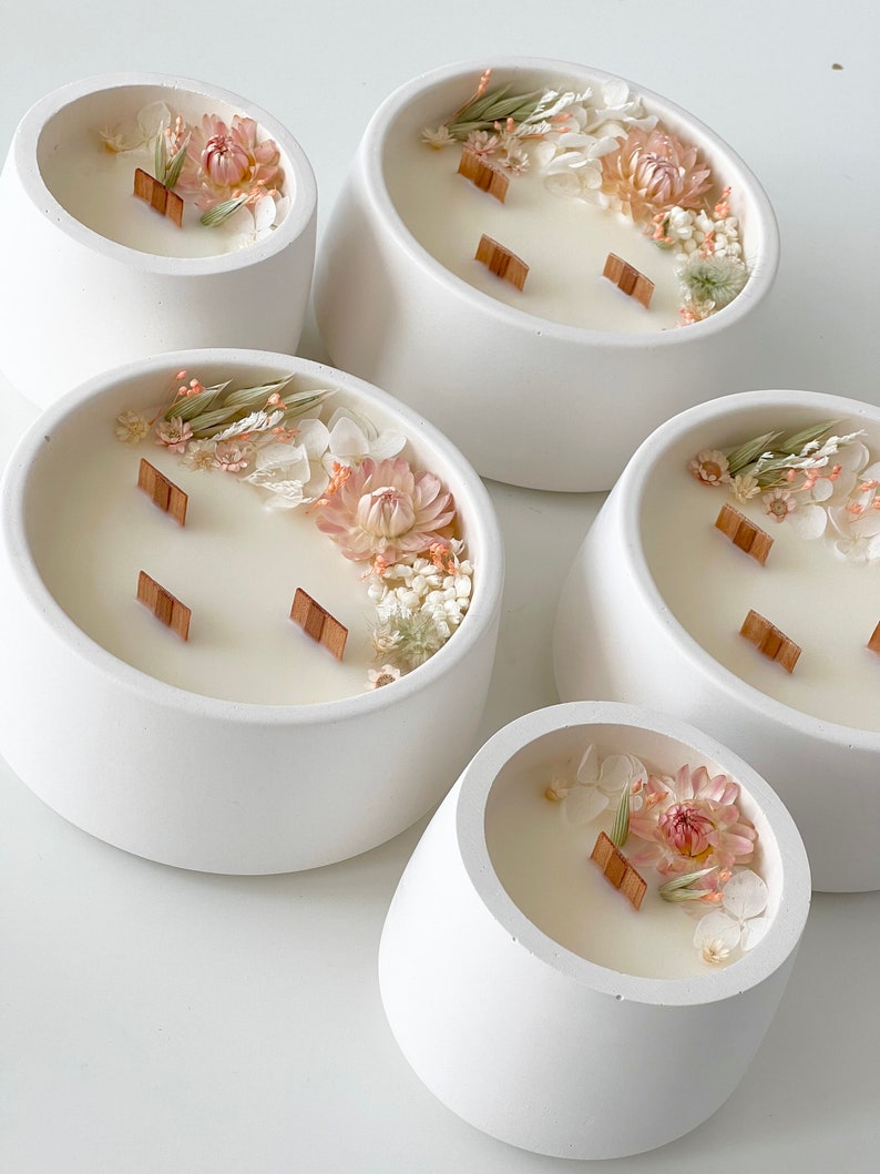 May include: Five white ceramic candle holders with a rounded, bowl-like shape. Each holder features a different arrangement of dried flowers and greenery, including white, pink, and green tones. The candles have wooden wicks.
