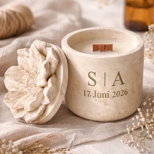 Könnte beinhalten: Eine beige Kerze in einem runden Betonbehälter mit einem Holzdocht. Der Behälter trägt die Initialen "S | A" und das Datum "17. Juni 2026". Ein dekorativer blütenförmiger Deckel liegt neben der Kerze. Die Kerze steht auf einer beigen Leinenoberfläche.