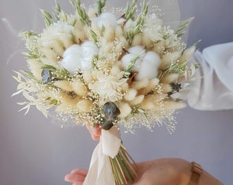 May include: A bouquet of dried flowers featuring fluffy white cotton, beige bunny tails, and delicate white blossoms. The stems are bound with a cream-colored ribbon. The arrangement is held in a person's hand.