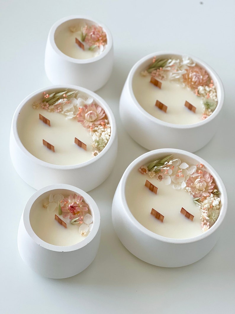 May include: Four white ceramic candle holders with a variety of dried flowers and wooden wicks. The candles are in different sizes.