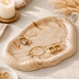 Könnte beinhalten: Ein beige-braun marmorierter Dekoteller mit Goldschmuck. Der Schmuck umfasst einen Ring mit einem weißen Stein, ein Armband mit Sternanhängern und zwei goldene Creolen. Eine brennende Kerze und Trockenblumen befinden sich im Hintergrund.