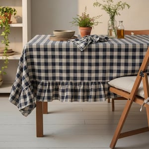 May include: A dining table with a navy and white gingham tablecloth and a frilled edge. A wooden chair is positioned beside the table. A shelf in the background displays pottery and plants. The table is set for a meal.