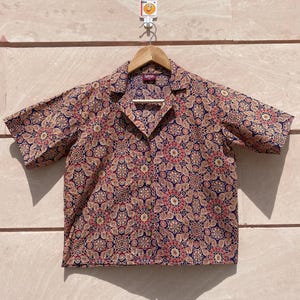 Könnte beinhalten: Ein kurzärmeliges Hemd mit einem Blumenmuster in Rot-, Beige- und Marineblautönen. Das Hemd hängt an einem Holzbügel. Das Hemd hat einen Kragen und Knöpfe.