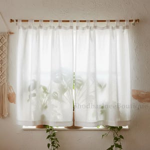 Puede incluir: Cortinas de lino blanco transparente colgadas de una barra de madera. Las cortinas son semitransparentes, permitiendo el paso de la luz. Se ve una planta detrás de las cortinas. Un tratamiento de ventana sencillo y elegante.