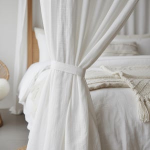 Può includere: Tende bianche per letto a baldacchino legate con una fascia in tessuto abbinata. Le tende sono realizzate in un tessuto leggero e strutturato. Il letto è coperto da biancheria da letto bianca e una coperta color crema.