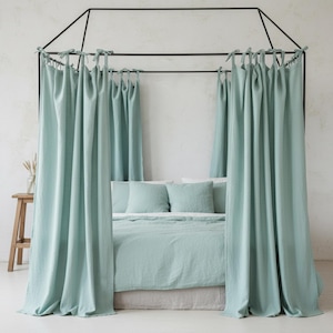 Pode incluir: Uma cama com edredão, almofadas e cortinas de linho verde menta. As cortinas estão amarradas a uma estrutura de dossel de metal preto. Um pequeno banco de madeira e um vaso com plantas secas são visíveis.