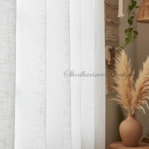May include: White sheer curtains with a subtle textured pattern. The curtains are hanging in front of a window, with a glimpse of a decorative vase with dried plants and the text "Shodharinee Boutique".