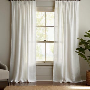 May include: Two white linen curtains hang on a dark brown rod, framing a window with a view of trees. The curtains are floor-length and slightly sheer, allowing natural light to filter through. A neutral-toned chair and a potted plant in a woven basket are visible.
