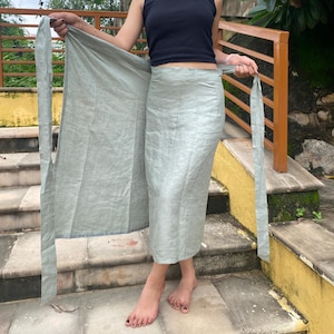 Peut inclure: Une jupe portefeuille vert clair avec de longues attaches, fabriquée dans un tissu léger. La jupe est présentée ouverte, révélant toute sa longueur et son design. La jupe est de couleur unie et semble faite à la main.