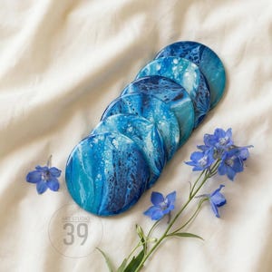 Può includere: Un set di sei sottobicchieri rotondi con un design astratto blu e bianco, disposti su un tessuto bianco. I sottobicchieri presentano un effetto marmorizzato, con sfumature di blu. Fiori blu e il testo "ART STUDIO 39" sono visibili.