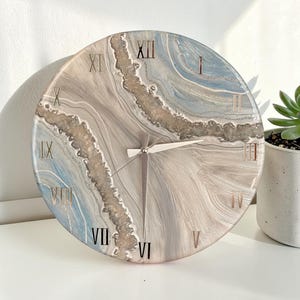 May include: Round wall clock with a marbled design in shades of blue, gray, and beige. The clock features gold Roman numerals and hands. The clock is a decorative home decor item.