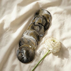 Può includere: Un set di sette sottobicchieri rotondi con un design marmorizzato in nero, oro e bianco. I sottobicchieri sono disposti su un tessuto color crema, con un fiore bianco e il testo "ART STUDIO 39" in basso a destra.