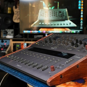 May include: A black audio mixing console with a wooden base. The console has multiple knobs, buttons, and sliders. The console is sitting on a wooden table.