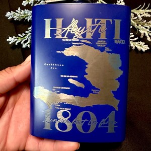 Könnte beinhalten: Eine blaue Flasche mit einer silberfarbenen Karte von Haiti und dem Text "Haiti Ayiti 1804". Die Flasche hat einen silbernen Verschluss und wird in einer Hand gehalten. Die Flasche hat eine rechteckige Form.