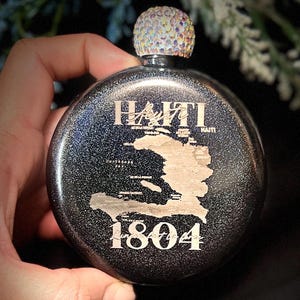 Könnte beinhalten: Eine runde, schwarze Flasche mit glitzerndem Finish. Die Flasche zeigt das Wort "Haiti" und eine goldfarbene Umrisskarte von Haiti. Das Jahr "1804" ist darunter aufgedruckt. Die Flasche hat einen dekorativen, juwelenbesetzten Verschluss.