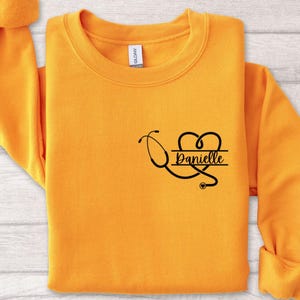 May include: A yellow sweatshirt with a black stethoscope and heart design. The name "Danielle" is written on a banner inside the heart.