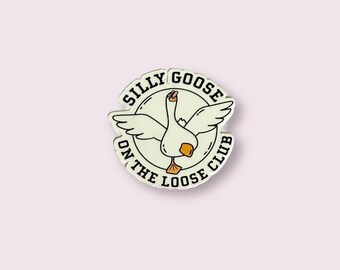 Puede incluir: Pin de esmalte blanco con un contorno negro de un ganso con las alas extendidas. El ganso está dentro de un círculo con el texto "Silly Goose On The Loose Club".