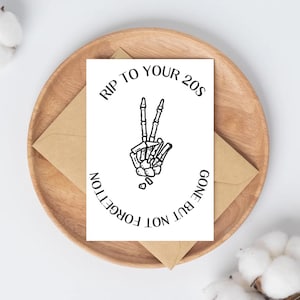 Könnte beinhalten: Eine weiße Grußkarte mit dem Text "RIP TO YOUR 20S GONE BUT NOT FORGOTTEN" und einer schwarzen Skeletthand, die ein Friedenszeichen zeigt. Die Karte liegt auf einem Holztablett mit einem braunen Umschlag.