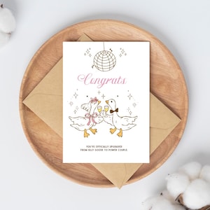 May include: A greeting card with the word "Congrats" in pink script, featuring two cartoon geese toasting champagne. The card is on a wooden tray with a brown envelope. Text reads: "You've officially upgraded from silly goose to power couple."