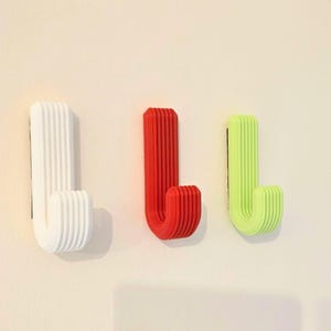 May include: Three J-shaped wall hooks mounted in a row. The hooks are white, red, and lime green, each featuring a distinct ribbed or layered texture along the vertical and curved surfaces, providing a uniform design for hanging various items.