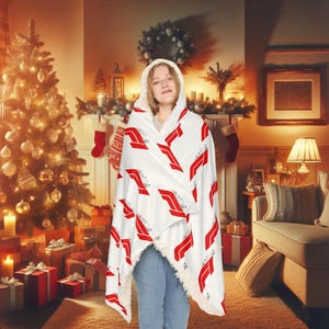 Puede incluir: Una manta blanca con capucha con un patrón geométrico rojo y blanco. La manta está drapeada sobre una persona en un ambiente acogedor con un árbol de Navidad y una chimenea.