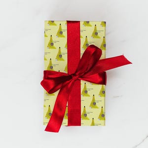 May include: A wrapped gift with yellow paper featuring a repeating pattern of a banana costume with the text "I've never looked so good." The gift is tied with a bright red satin ribbon and bow.