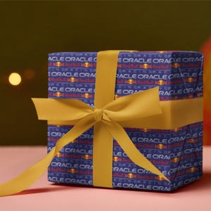 May include: A wrapped gift box with a blue and red patterned design, featuring the text "ORACLE RACING". The box is tied with a yellow ribbon and bow, set against a pink surface.