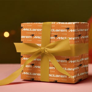 Puede incluir: Una caja de regalo envuelta en papel naranja con el logotipo y texto de McLaren Formula 1 Team. La caja está atada con una cinta y lazo amarillo mostaza. Fondo rosa.