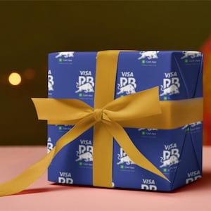 Può includere: Un pacco regalo avvolto in carta blu con il logo Visa PR, legato con un nastro dorato. La scatola è su una superficie rosa, con elementi sfocati sullo sfondo.