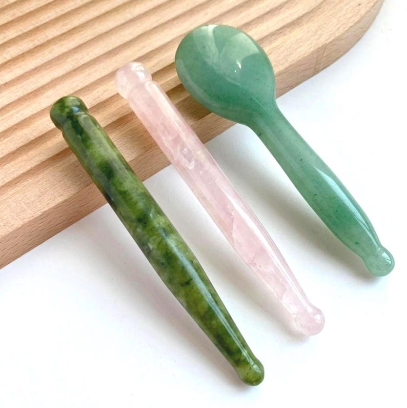 May include: Three smooth, polished stone massage tools. One is a dark green mottled cylindrical wand, one is a translucent pale pink cylindrical wand, and one is a light green rounded spoon-shaped tool with a long, tapered handle.
