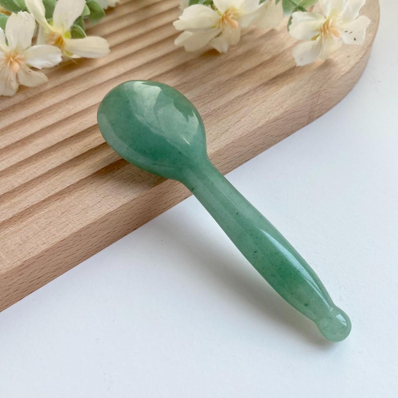May include: A smooth, polished green stone tool shaped like a small spoon with a rounded bowl and a tapered handle that ends in a small, rounded knob. The surface of the stone features natural variations in color and texture throughout the piece.