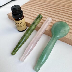 May include: A small brown glass bottle of facial oil with a black cap, a dark green stone massage wand, a pale pink stone massage wand, and a light green stone massage tool with a rounded head, all arranged on a white surface.