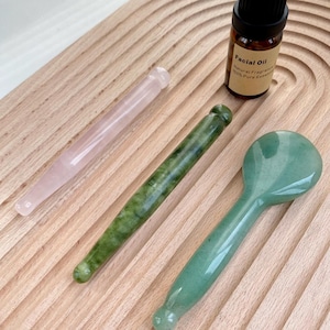 May include: A light pink quartz massage wand, a dark green jade massage wand, a smooth light green stone facial tool with a rounded head, and a small brown glass bottle labeled Facial Oil with a black cap, all arranged on a ridged surface.