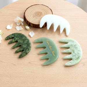 Puede incluir: Una colección de peines de jade en varios tonos de verde y blanco, dispuestos sobre una superficie de madera. Un peine tiene el texto "Calm Aura Haven" en dorado. Los peines tienen forma curva con múltiples dientes, diseñados para el cuidado del cabello o del cuero cabelludo.