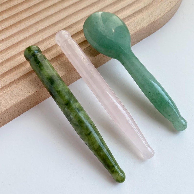 May include: Three smooth, polished stone massage tools. One is a dark green mottled cylindrical wand, one is a pale translucent pink cylindrical wand, and one is a light green spoon-shaped tool with a rounded head and a tapered handle.