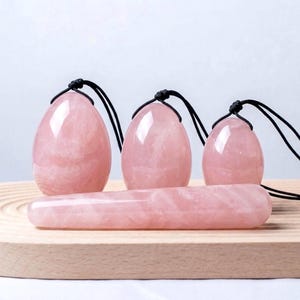 以下が含まれることがあります： 4つのピンク色のローズクォーツアイテム：黒いコードが付いた卵型のペンダント3つと、長く丸みを帯びたワンド。明るい木製の表面にアイテムが展示されています。