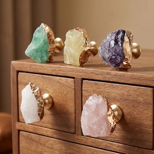 May include: Five cabinet knobs featuring raw, irregular crystal stones in green, yellow, purple, white, and pink. Each stone is accented with a textured gold-toned metal edge and a smooth, round gold-toned metal base for mounting to furniture.