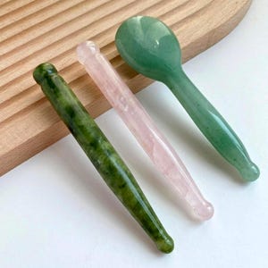 Puede incluir: Tres herramientas faciales de piedras preciosas: un rodillo de jade verde, un rodillo de cuarzo rosa y una herramienta en forma de cuchara de jade verde. Las herramientas están dispuestas sobre una superficie de madera clara, mostrando sus superficies pulidas y colores naturales.