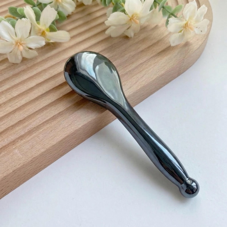 May include: A smooth, dark gray, metallic-looking massage tool with a rounded, spoon-shaped head and a tapered handle that ends in a small, spherical knob. The surface is highly reflective, showing light highlights across its curved, solid form.