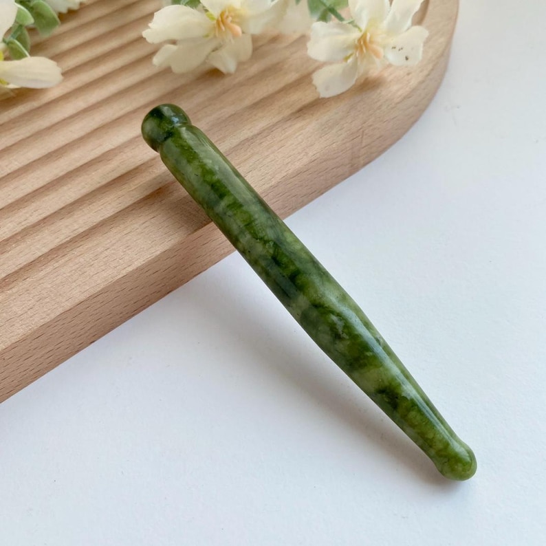 May include: A smooth, cylindrical massage wand crafted from dark green, mottled stone. The tool features a rounded, bulbous tip at one end and a slightly tapered, rounded end at the other, showcasing natural variations in the deep green mineral color.