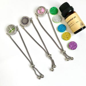 Könnte beinhalten: Drei silberne Kettenarmbänder mit jeweils einem runden Anhänger. Die Anhänger zeigen einen Lebensbaum, ein Mandala und ein Katzenmotiv. Die Armbänder sind mit einem Schiebekugel verstellbar. Das Bild zeigt außerdem eine kleine Flasche ätherisches Öl und fünf bunte runde Pads.