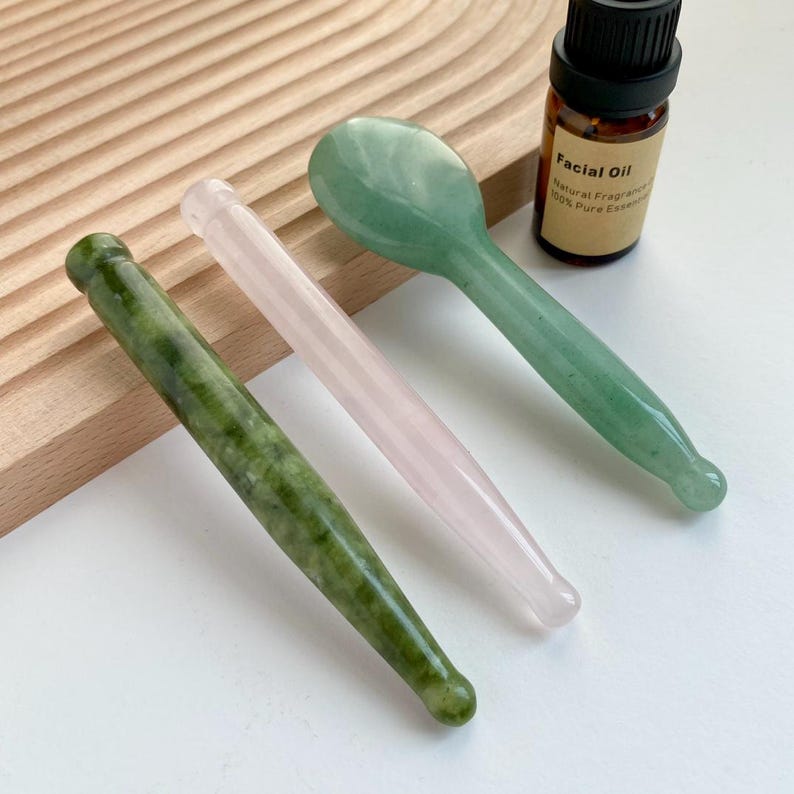 May include: A dark green stone massage wand, a light pink stone massage wand, and a green stone facial spoon next to a small brown glass bottle labeled Facial Oil with a black cap. The items are arranged on a white surface.