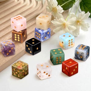 May include: A selection of gemstone dice in various colours, including pink, brown, black, blue, green, and red. Each die features gold-coloured dots. The dice are displayed on a wooden surface with white flowers in the background.