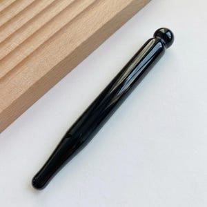 May include: A smooth, polished black massage wand with a cylindrical body and a rounded spherical tip at one end. The tool features a glossy, reflective surface and tapers slightly toward the opposite end, presenting a sleek, solid black appearance.