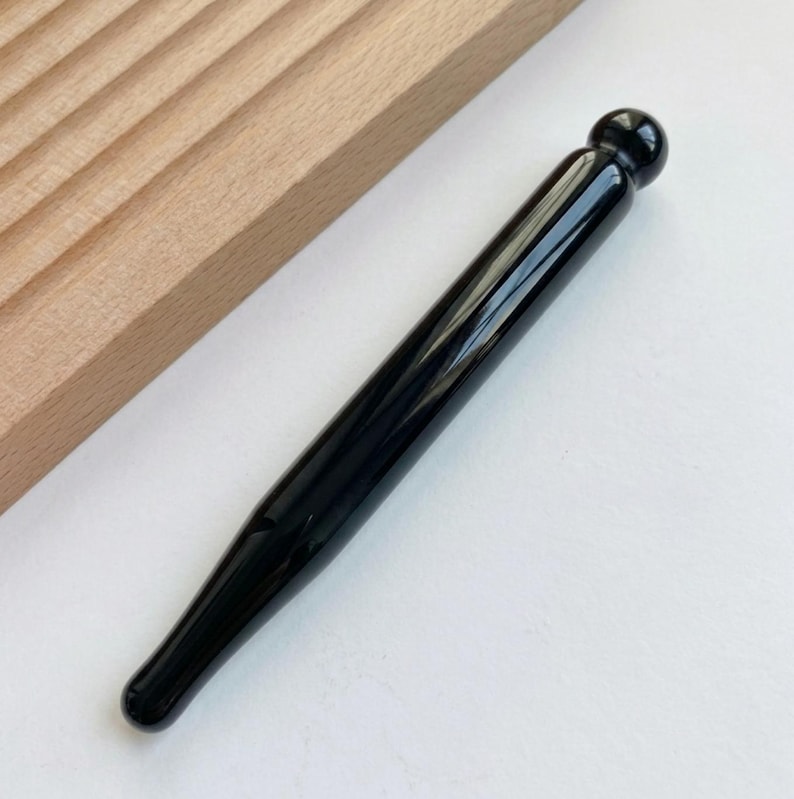 May include: A smooth, polished black massage wand with a cylindrical body and a rounded spherical tip at one end. The tool features a glossy, reflective surface and tapers slightly toward the opposite end, presenting a sleek, solid black appearance.